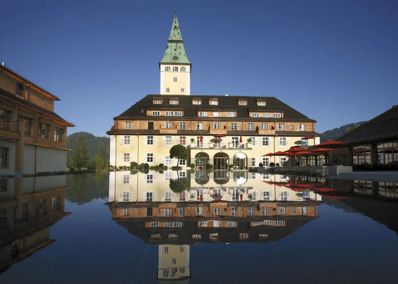 Majestätisches Schloss Elmau mit Turm und perfekter Spiegelung im Wasserbecken unter blauem Himmel.