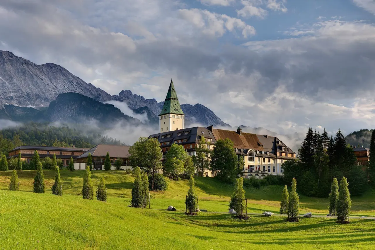 Schloss Elmau Hotel in den bayerischen Alpen mit majestätischem Bergpanorama und grünen Wiesen unter bewölktem Himmel.
