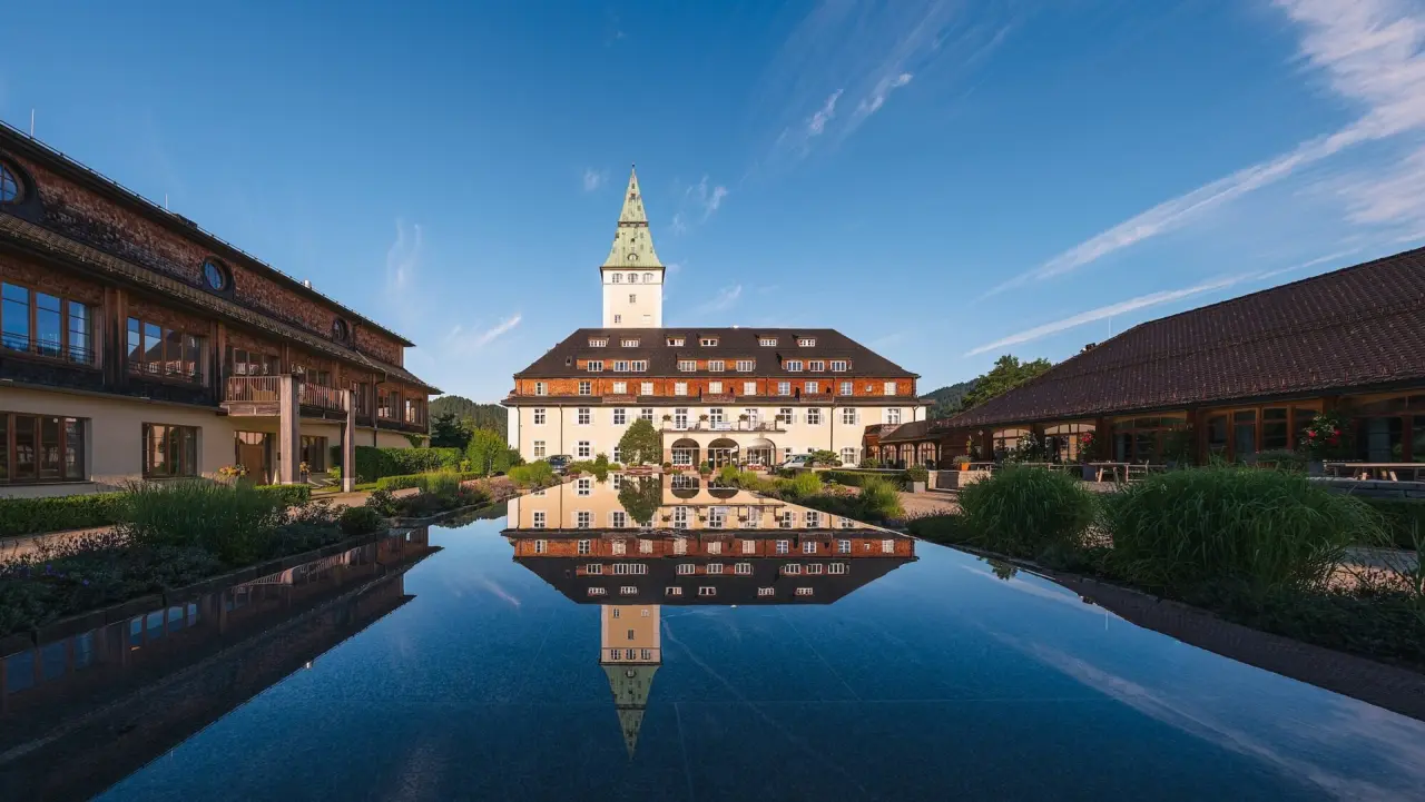 Majestätisches Schloss Elmau Hotel mit Turm und perfekter Spiegelung im Wasserbecken unter blauem Himmel.
