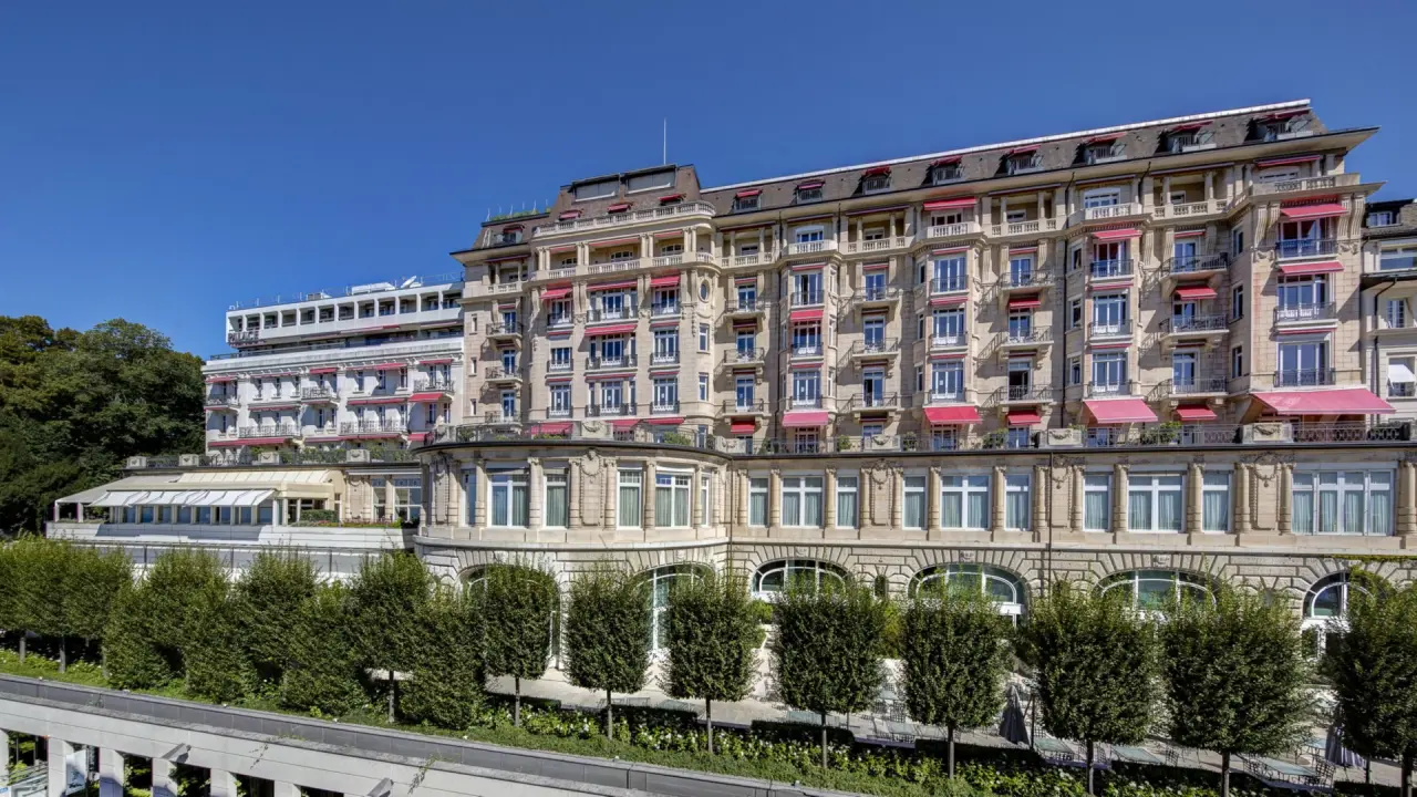 Prächtige Aussenansicht des historischen Hotel Lausanne Palace mit roten Markisen und grünen Bäumen unter blauem Himmel.