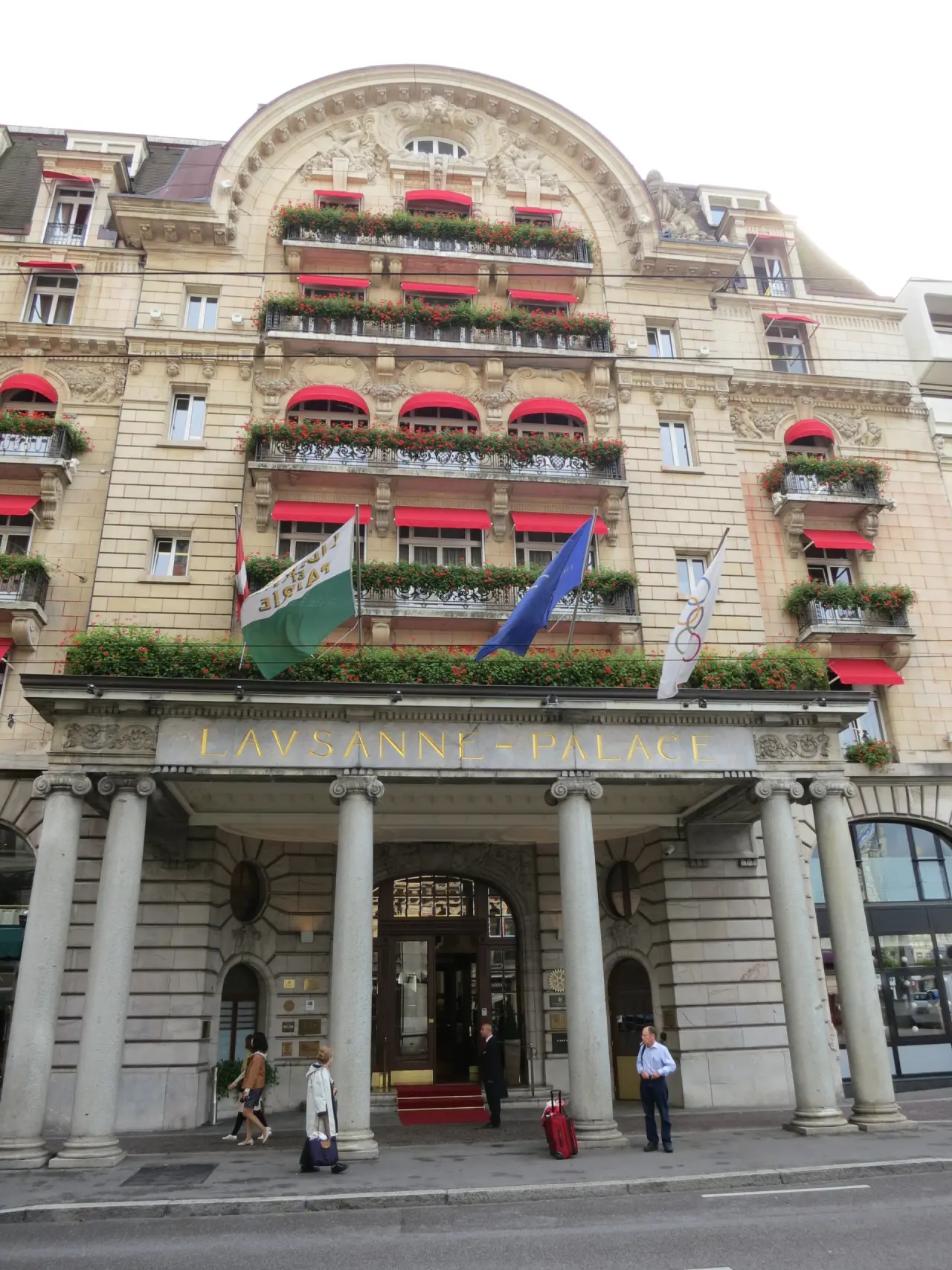 Prächtige Fassade des Hotel Lausanne Palace mit roten Markisen, blühenden Balkonen und imposantem Säuleneingang.
