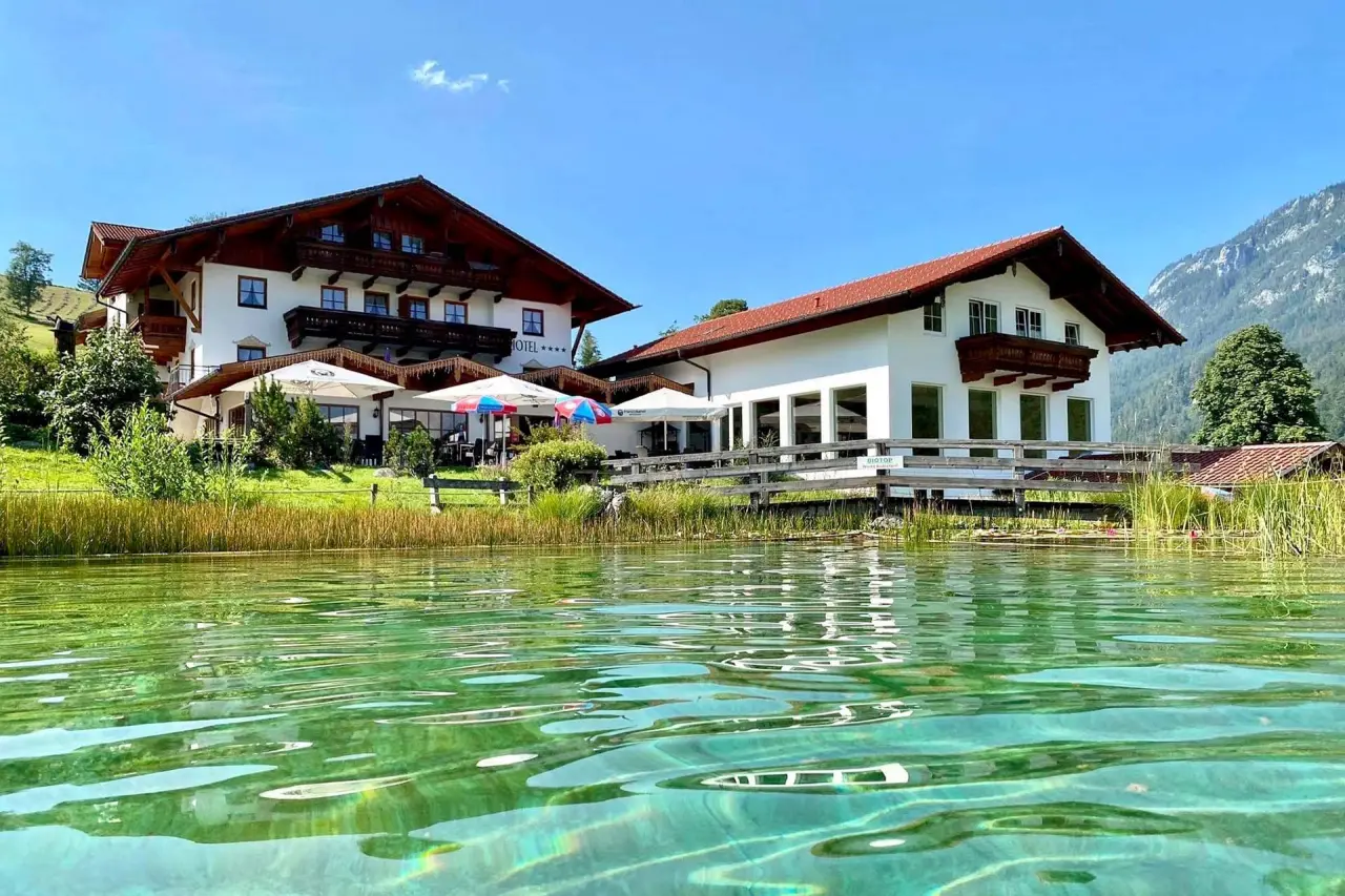 Naturhotel Reissenlehen mit traditioneller Architektur am idyllischen Biotop-Teich und Bergkulisse unter blauem Himmel.