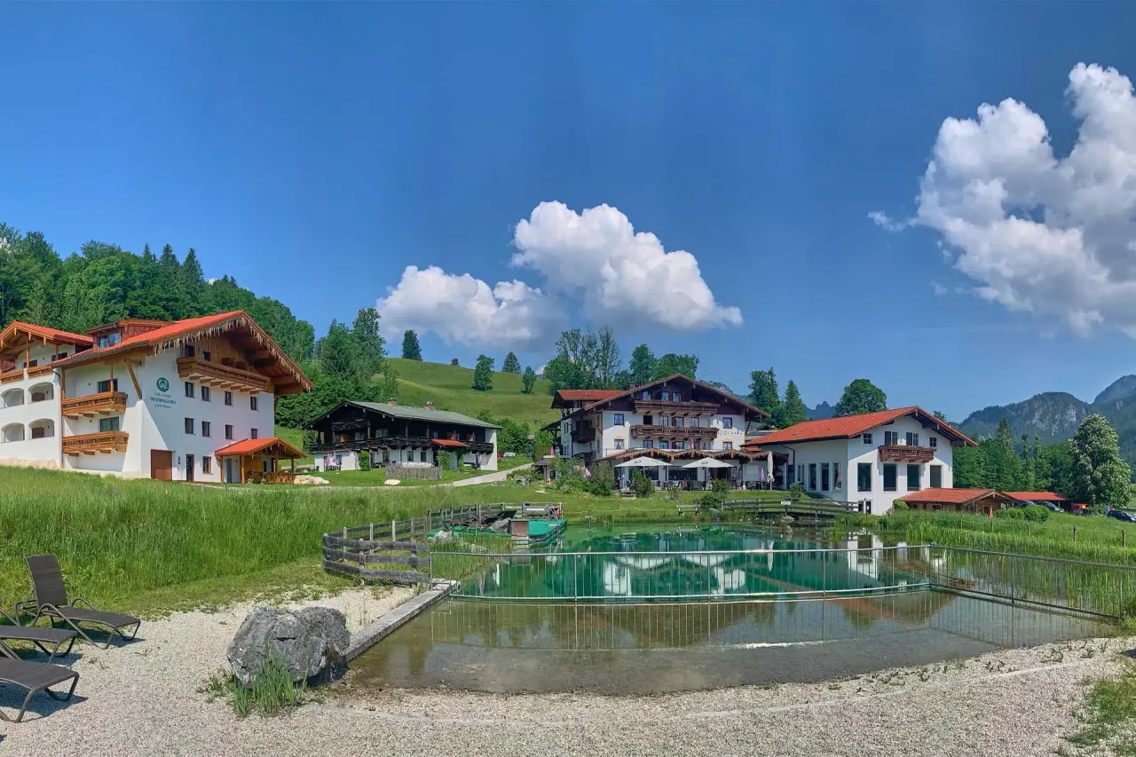 Panoramaansicht des Naturhotel Reissenlehen mit Natur-Badeteich, Liegewiese und Alpenkulisse unter blauem Himmel.
