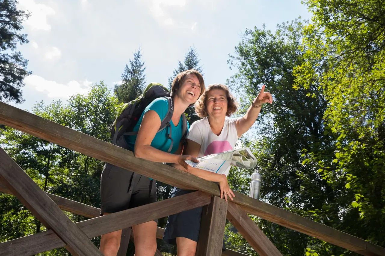 Fröhliche Wanderinnen mit Karte auf einer Holzbrücke im Wald, ideal für Outdoor-Erlebnisse rund um das Wellness-Hotel Cafe Talblick.