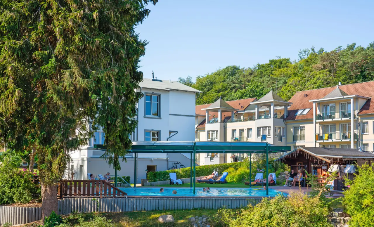 Gepflegter Aussenpool mit Gästen und Sonnenliegen, umgeben von Hotelvillen und Bäumen im Ostseehotel - Villen im Park.