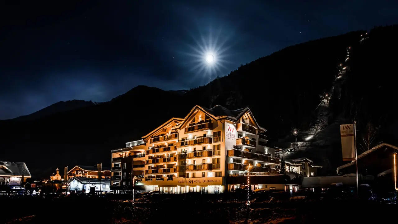 Beleuchtetes Hotel Mallaun bei Nacht im Winter mit Vollmond und verschneiter Bergkulisse. Perfekt für Wellness & Ski.