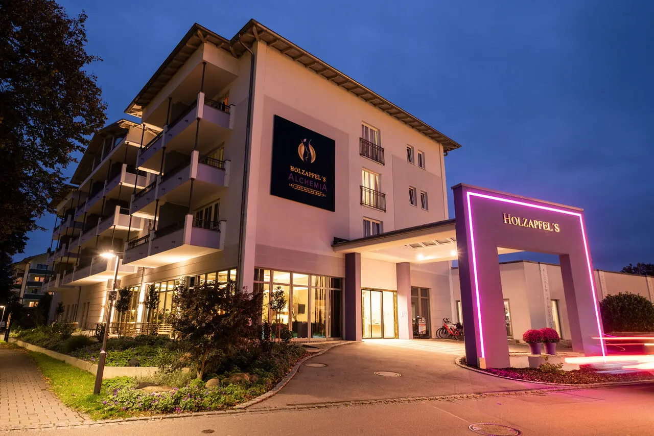 Moderne Aussenansicht des Holzapfel's Alchemia Spa- und Wellnesshotels bei Nacht mit beleuchtetem Eingang und Neon-Bogen.