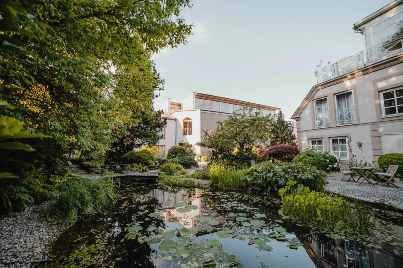 Idyllischer Hotelgarten mit Teich, Seerosen und Hotelgebäuden im Garten-Hotel Ochensberger.