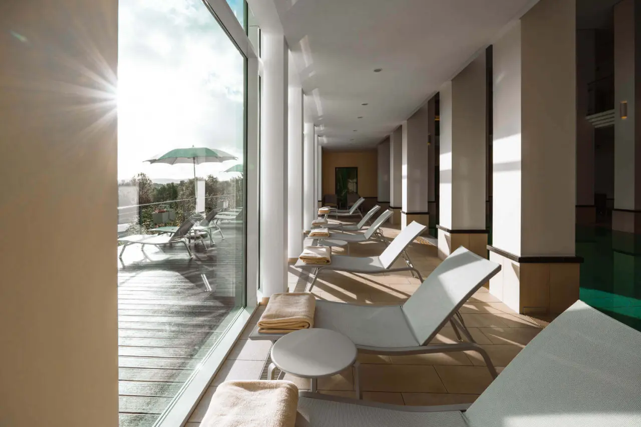 Heller Entspannungsbereich mit Liegestühlen und Blick auf die Sonnenterrasse am Innenpool im DIEDRICH Wellnesshotel & SPA.