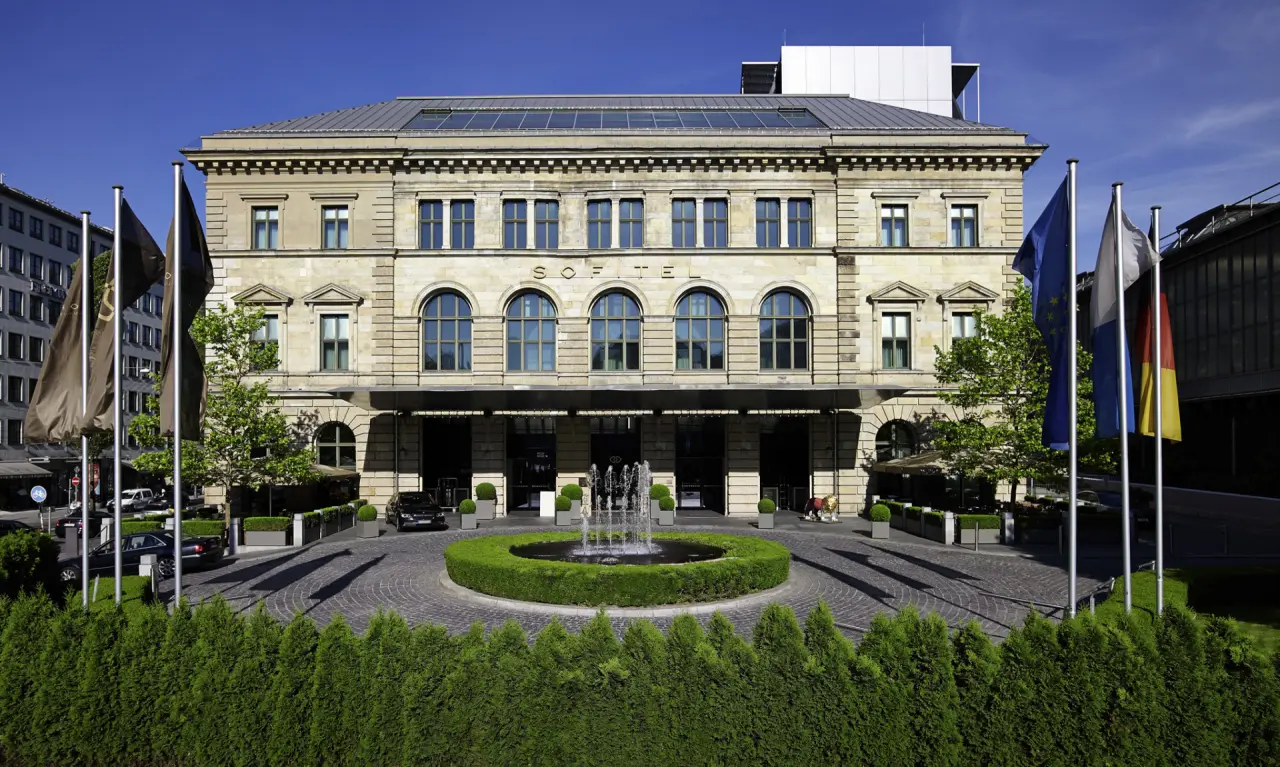 Elegante Aussenansicht des Sofitel Munich Bayerpost mit historischer Fassade, Brunnen und gepflegter Zufahrt in München.