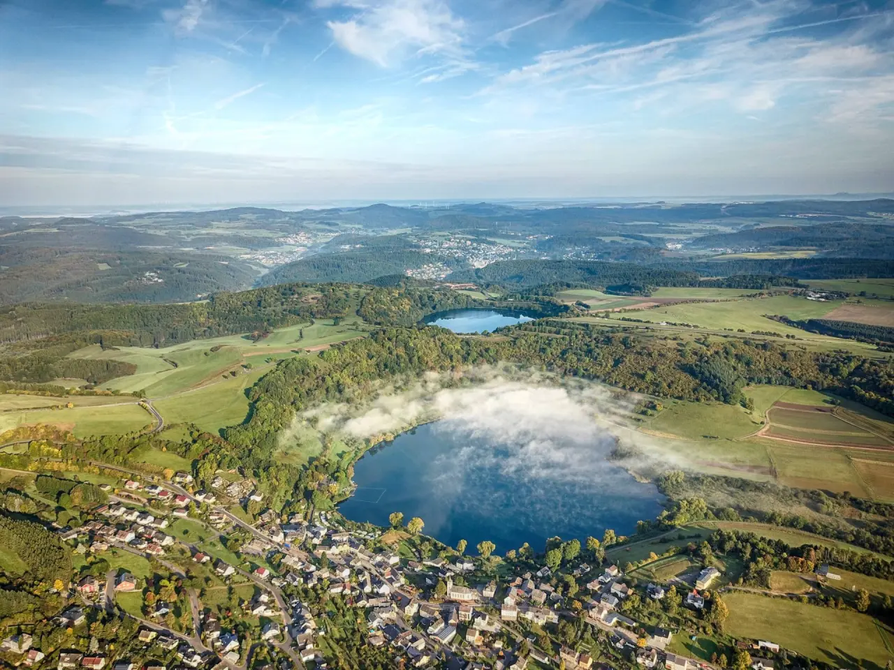 Luftaufnahme der malerischen Seenlandschaft mit Nebelschwaden und Dorf um Michels Wohlfühlhotel.