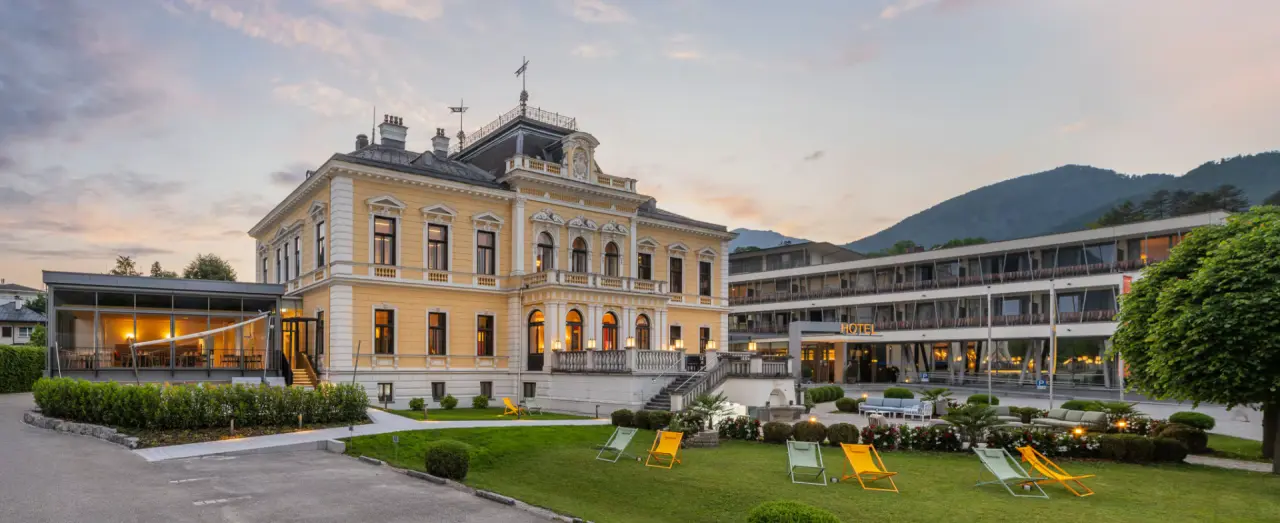 Abendansicht des Villa Seilern Vital Resort mit historischer Villa, modernem Anbau und Gartenbereich vor Bergkulisse.