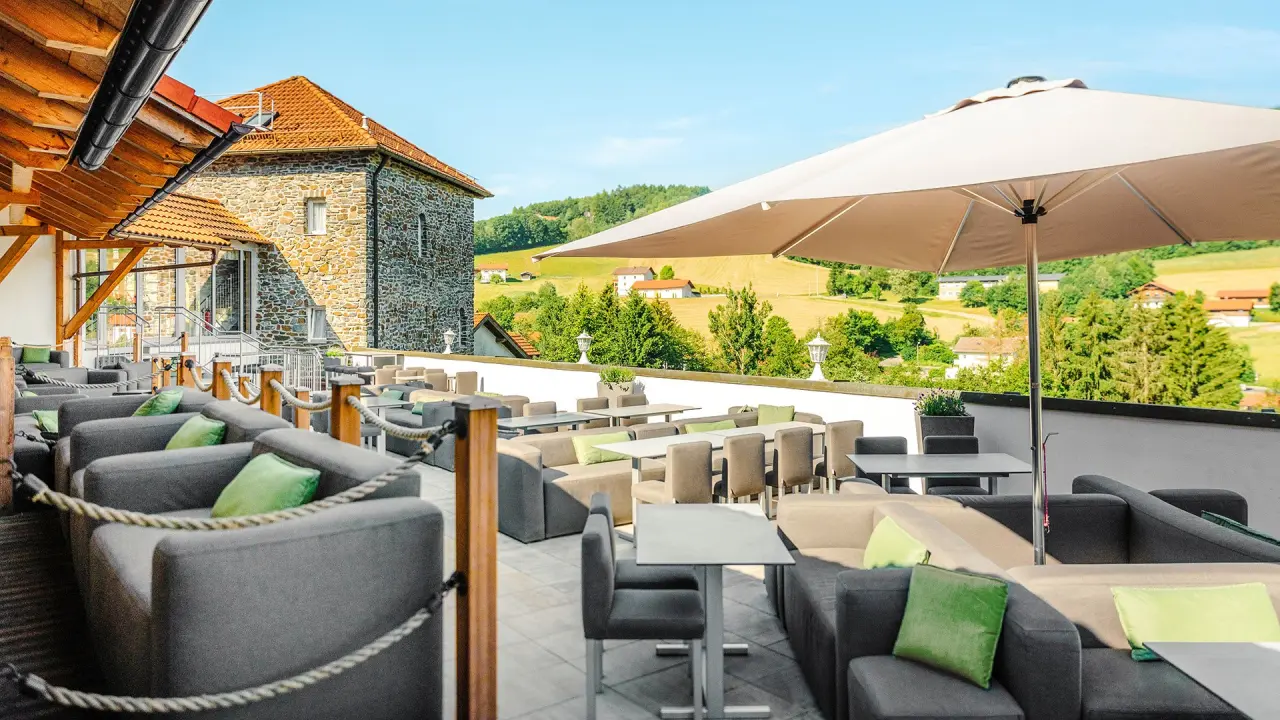Sonnige Restaurant-Terrasse mit bequemen Lounge-Möbeln und Panoramablick auf die bayerische Landschaft im Burghotel Am Hohen Bogen.
