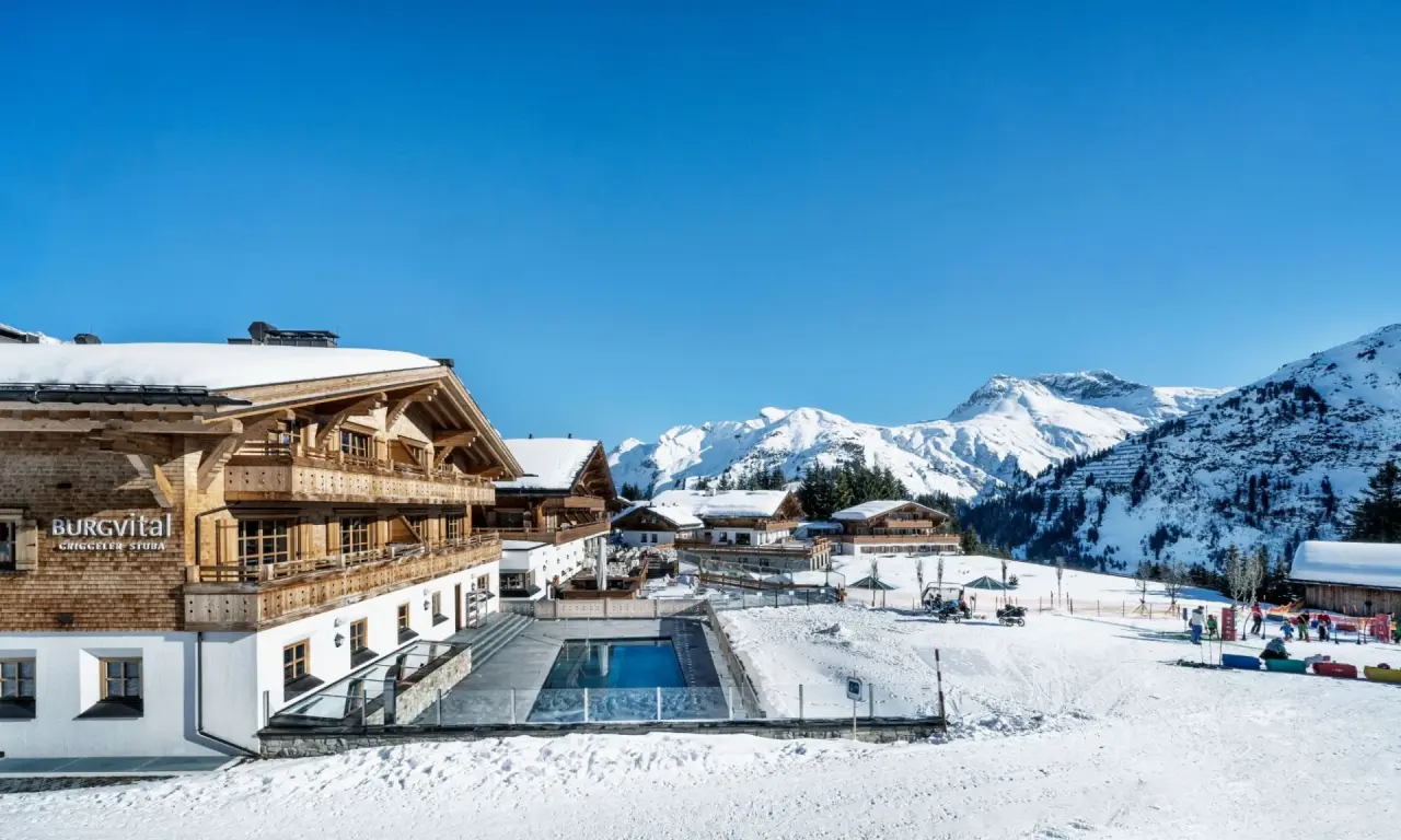Aussenansicht des Burg Hotel mit beheiztem Aussenpool und verschneiten Alpenbergen unter blauem Himmel im Winter.