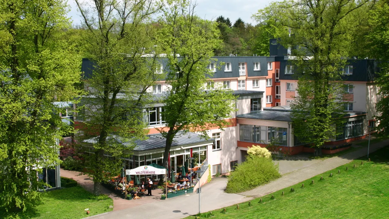 Aussenansicht des TRIHOTEL Rostock mit grünen Bäumen und der belebten Waldterrasse im Frühling.
