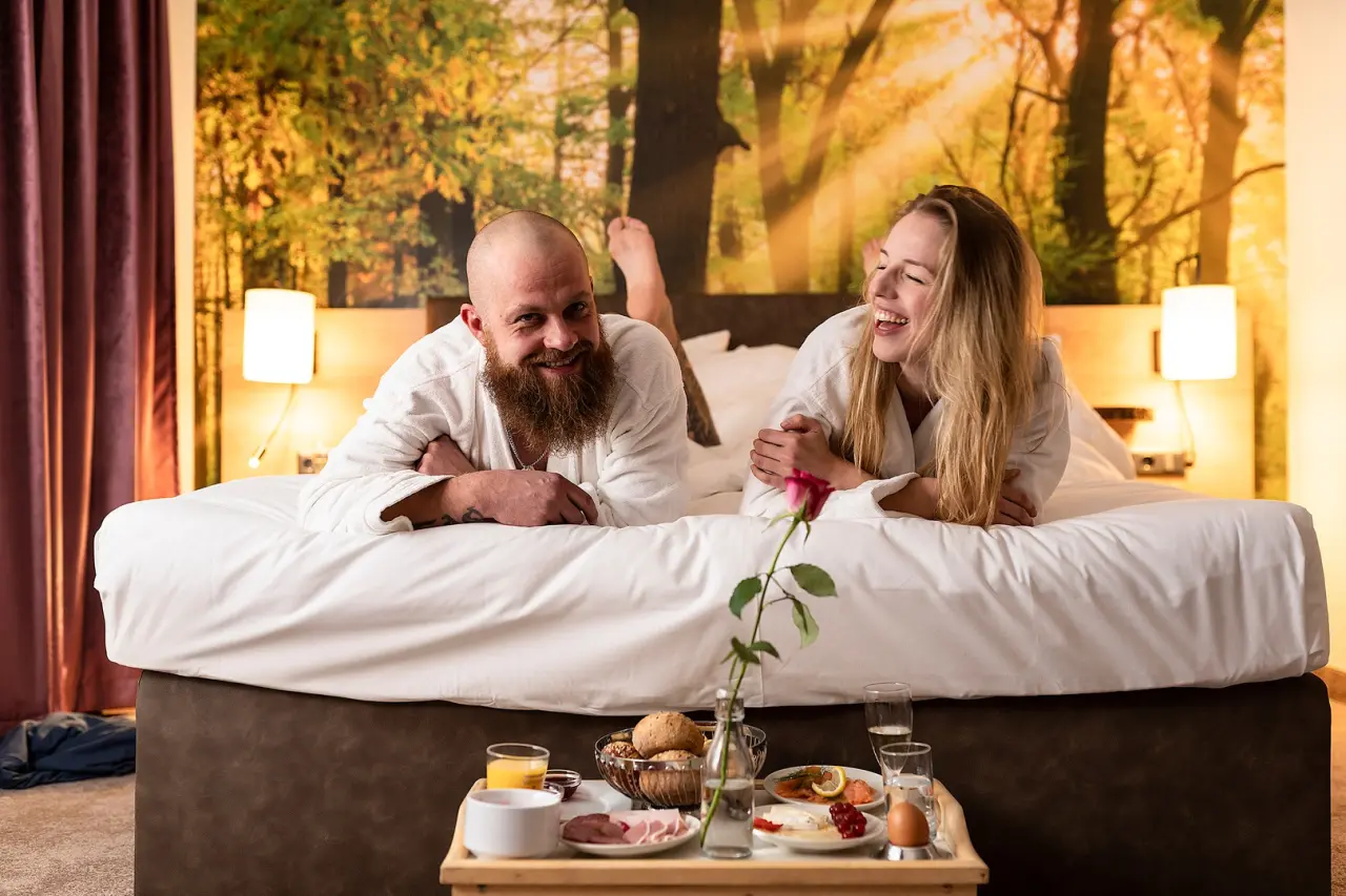 Glückliches Paar im Bademantel genießt Frühstück im Bett in einem komfortablen Hotelzimmer im TRIHOTEL Rostock.