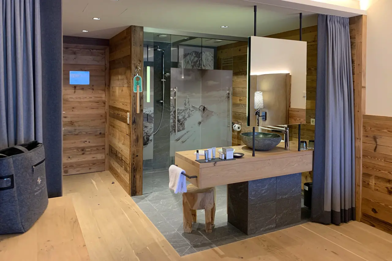 Modernes Badezimmer im Hotel Edelweiss mit Holzwaschtisch, Steinbecken und Glasdusche mit Skifahrer-Motiv.