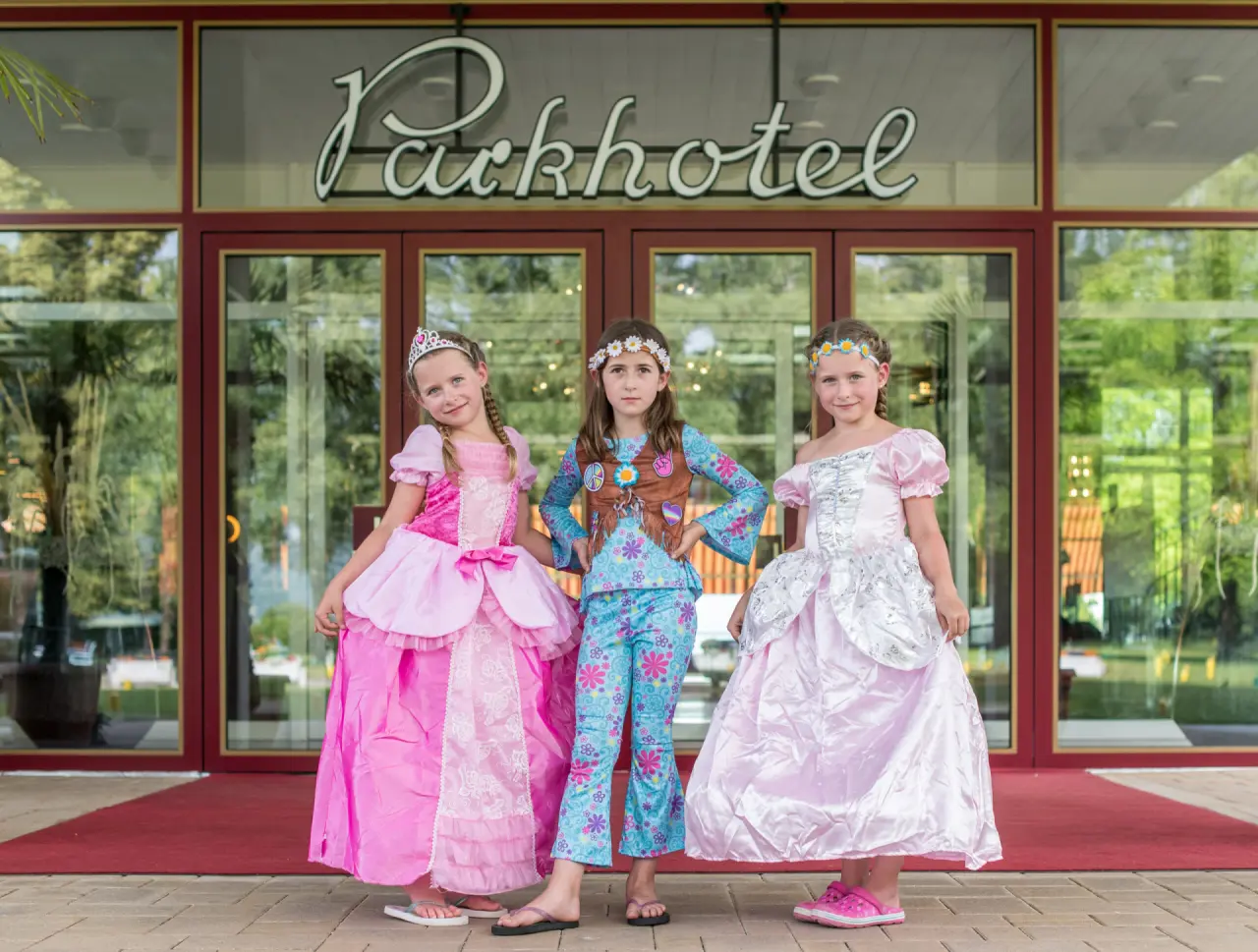 Drei Mädchen in bunten Kostümen vor dem Haupteingang des Parkhotel Pörtschach an einem sonnigen Tag.