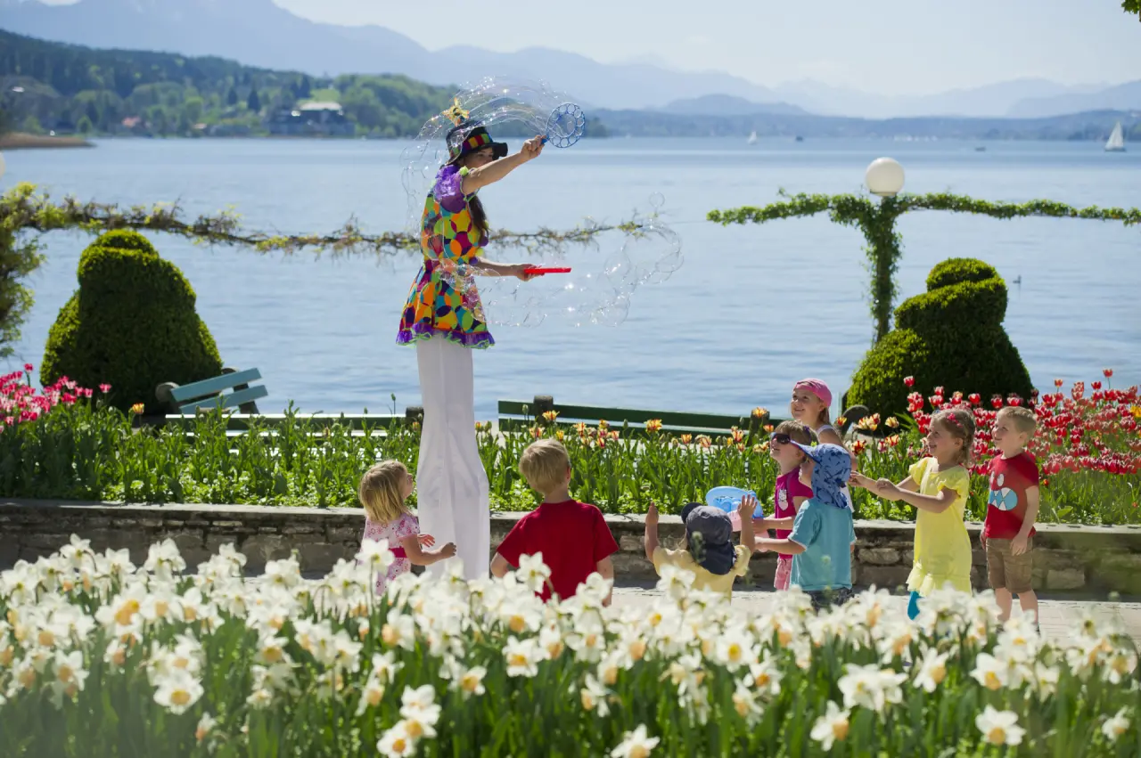 Fröhliche Kinder spielen mit Seifenblasen einer Stelzenläuferin im blühenden Garten des Parkhotel Pörtschach am Wörthersee.
