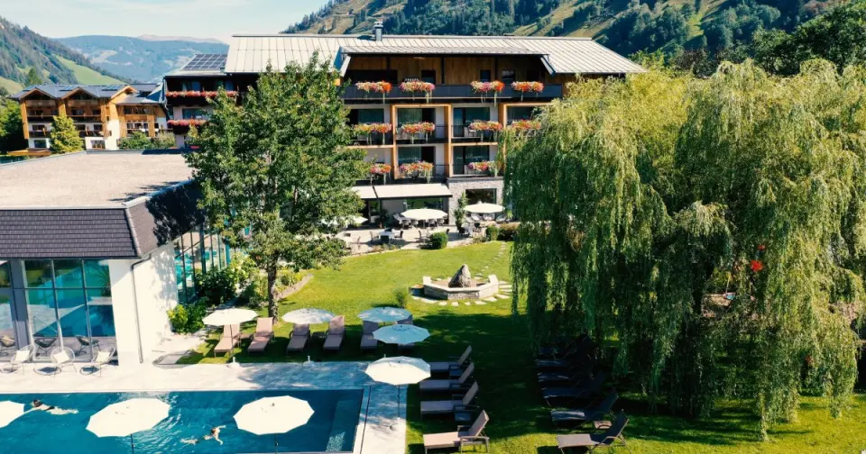 Aussenansicht des Hotel Rauriserhof mit sonnigem Garten, grossem Aussenpool und blühenden Balkonen vor Bergkulisse.