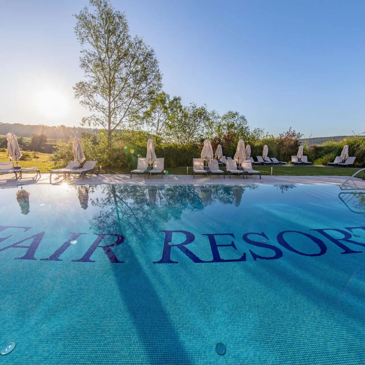 Aussenpool des Fair Resort bei Sonnenuntergang mit Liegestühlen und grüner Landschaft im Hintergrund.