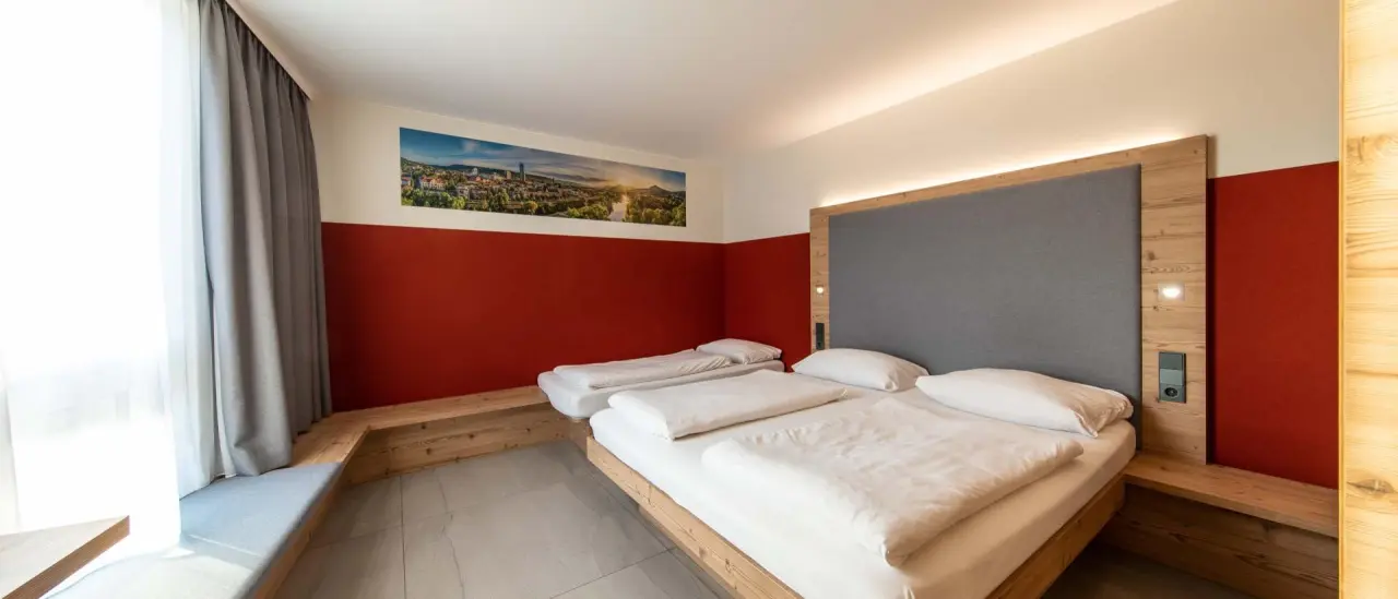 Modernes Doppelzimmer im Fair Hotel mit Holzelementen, roter Wand und Stadtpanorama-Bild über dem Bett.