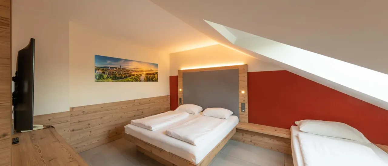 Gemütliches Doppelzimmer mit Holzelementen, roter Akzentwand und Zusatzbett im Fair Hotel.