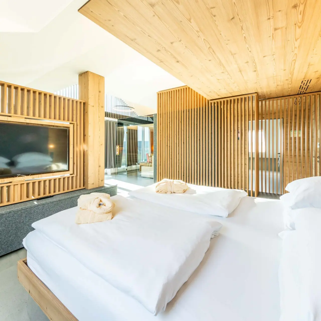Modernes Hotelzimmer im Fair Hotel mit hellem Holzdesign, Doppelbett, Bademänteln und Blick auf den Wohnbereich.