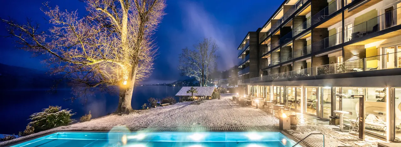 Winterliche Nachtansicht der Villa Postillion am See mit beleuchtetem Aussenpool, verschneitem Ufer und glitzerndem See.