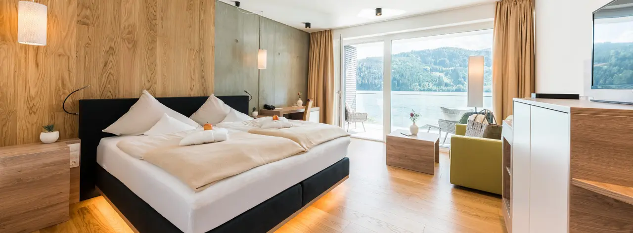 Modernes Hotelzimmer der Villa Postillion am See mit Doppelbett, Balkon und Panoramablick auf den Wörthersee und die Berge.