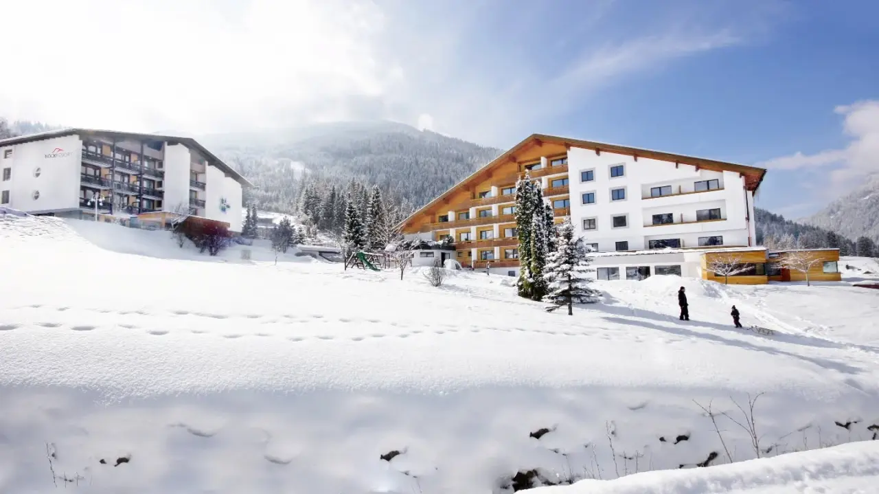 Winteransicht des Hotel NockResort mit modernen Gebäuden, umgeben von verschneiten Bergen und Bäumen unter blauem Himmel.