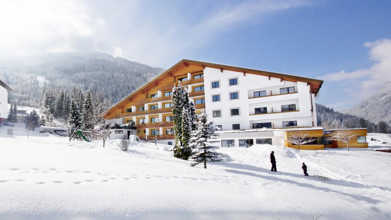Hotel NockResort in verschneiter Alpenlandschaft mit Tannen und spielenden Kindern im Vordergrund.