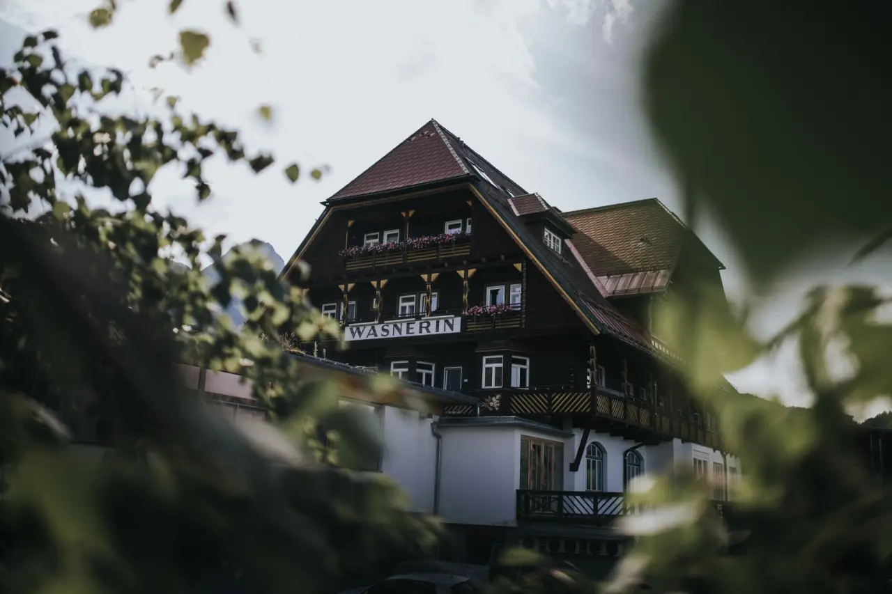 Traditionelles Hotel DIE WASNERIN mit Holzbalkonen und Blumenkästen, umgeben von grünen Bäumen unter hellem Himmel.