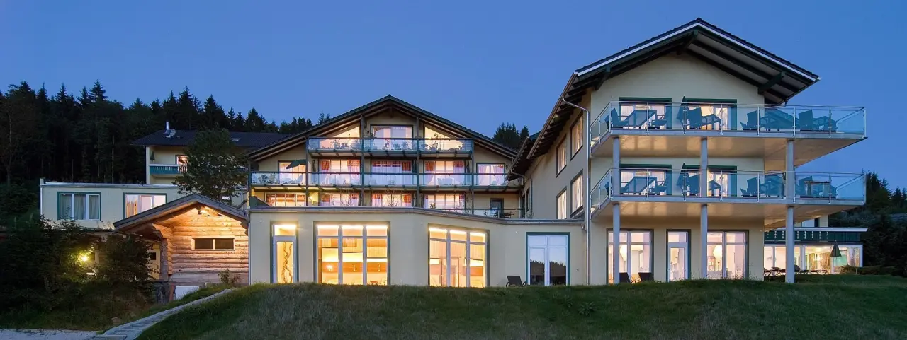 Aussenansicht des Hotel Reblinger Hof bei Dämmerung mit beleuchteten Balkonen und Wald im Hintergrund.