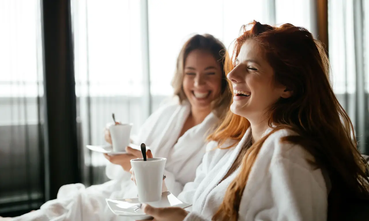 Zwei lachende Frauen in Bademänteln genießen Kaffee oder Tee im Romantik Hotel Haus Platte. Entspannung pur.