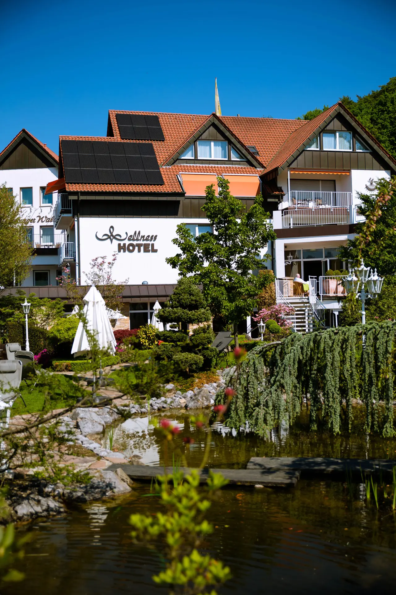 Aussenansicht des Ringhotel Teutoburger Wald mit 'Wellness Hotel' Schriftzug, Gartenanlage und Teich unter blauem Himmel.