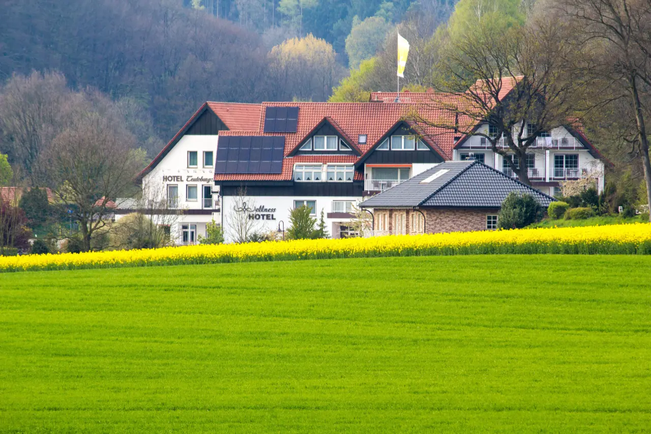 Aussenansicht des Ringhotel Teutoburger Wald mit gelbem Rapsfeld und grünen Wiesen im Vordergrund, umgeben von Wald.