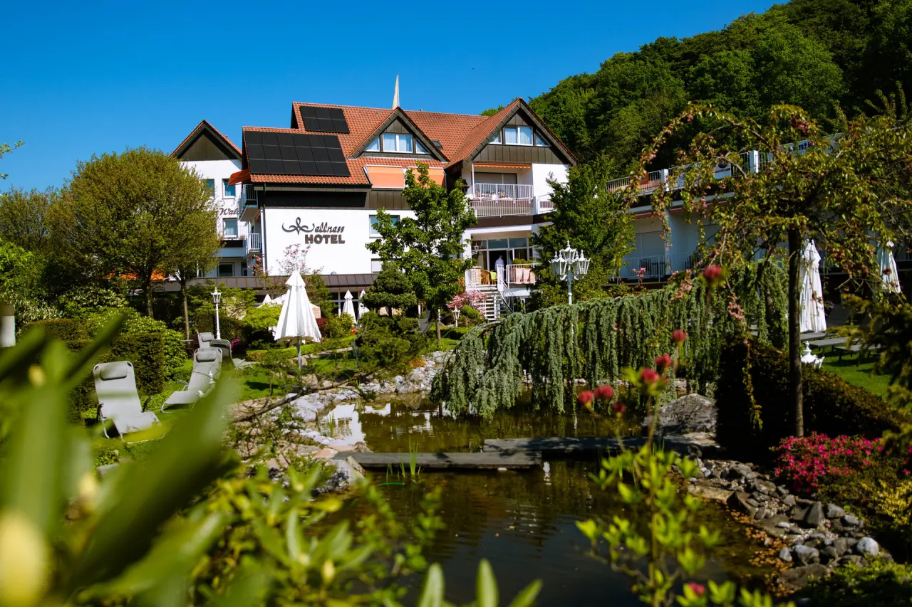 Aussenansicht des Ringhotel Teutoburger Wald mit Garten, Teich und 'Wellness HOTEL' Schriftzug unter blauem Himmel.