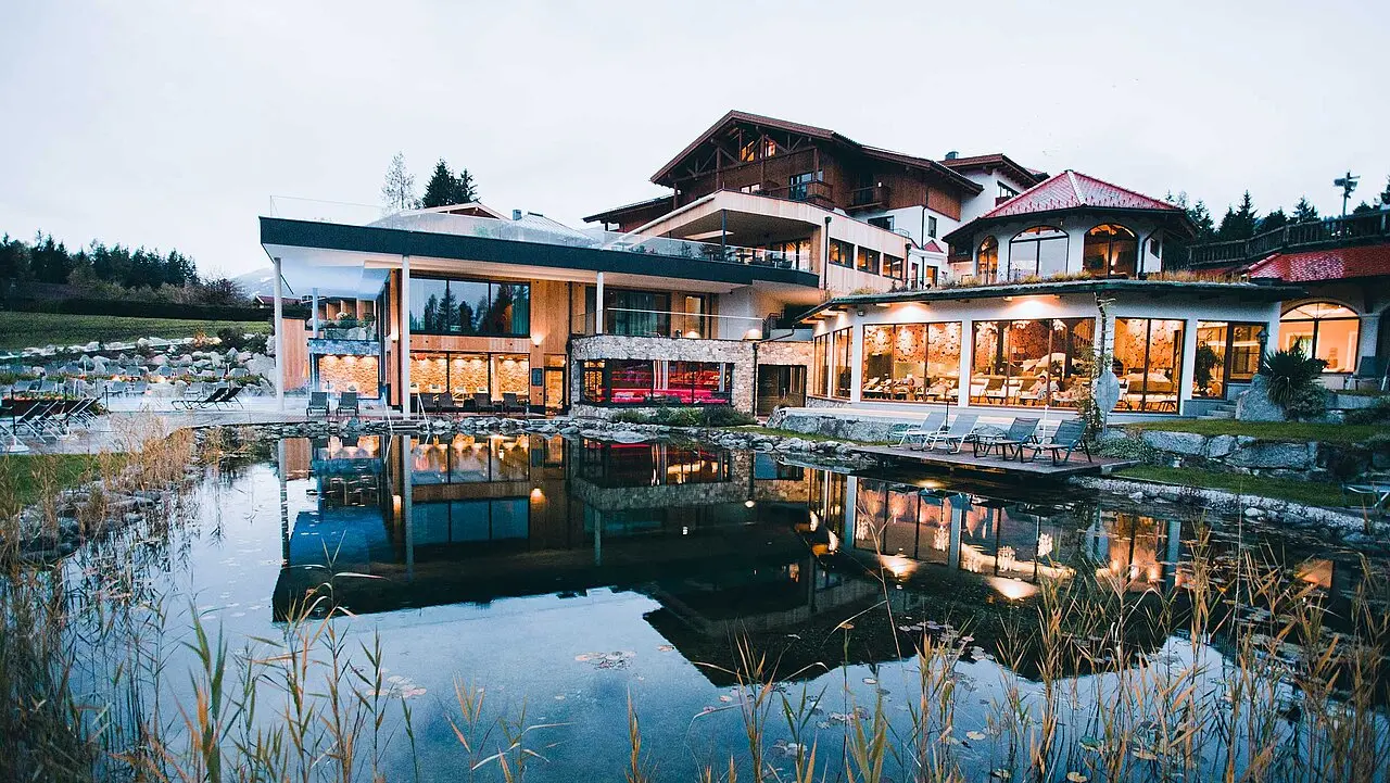 Abendansicht des Gut Sonnberghof Naturhotel mit beleuchteter Fassade und Spiegelung im Naturteich. Wellness-Oase in den Alpen.