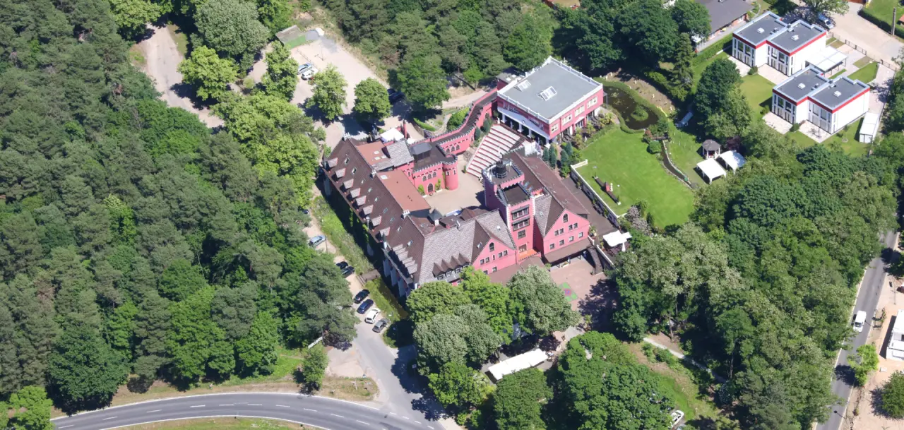Luftaufnahme des The Lakeside Burghotel zu Strausberg, ein malerisches rotes Burghotel inmitten von gruenen Waeldern.