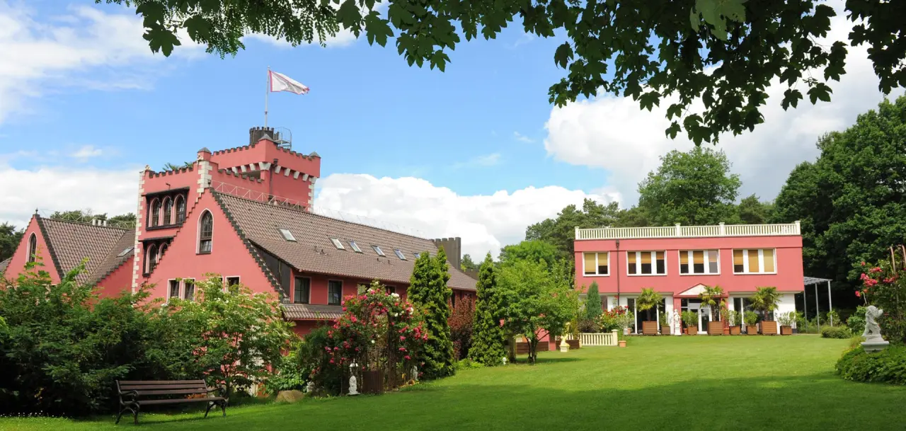 Das The Lakeside Burghotel zu Strausberg: Rosafarbenes Schlossgebäude mit Turm, grüner Gartenanlage und blühenden Rosen unter blauem Himmel.