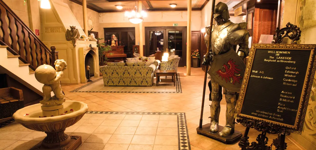 Historische Lobby des The Lakeside Burghotel zu Strausberg mit Ritterrüstung, Brunnen, Treppe und gemütlicher Sitzecke.