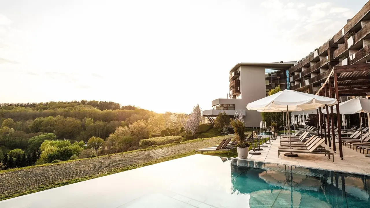 Aussenpool des Falkensteiner Balance Resort Stegersbach mit Liegen, Sonnenschirmen und Blick auf die grüne Landschaft.