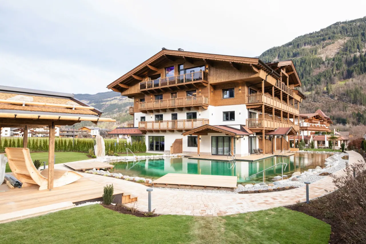 Aussenansicht des Hotel Wöscherhof mit traditioneller Holzarchitektur, umgeben von einem natürlichen Schwimmteich und Liegewiesen vor Bergkulisse.