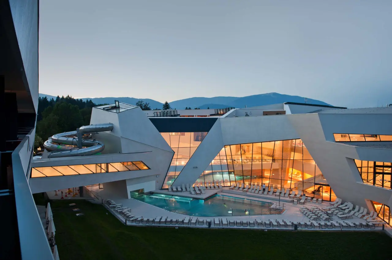 Modernes Thermenhotel Karawankenhof bei Dämmerung mit beleuchtetem Aussenpool, Wasserrutsche und Bergkulisse.