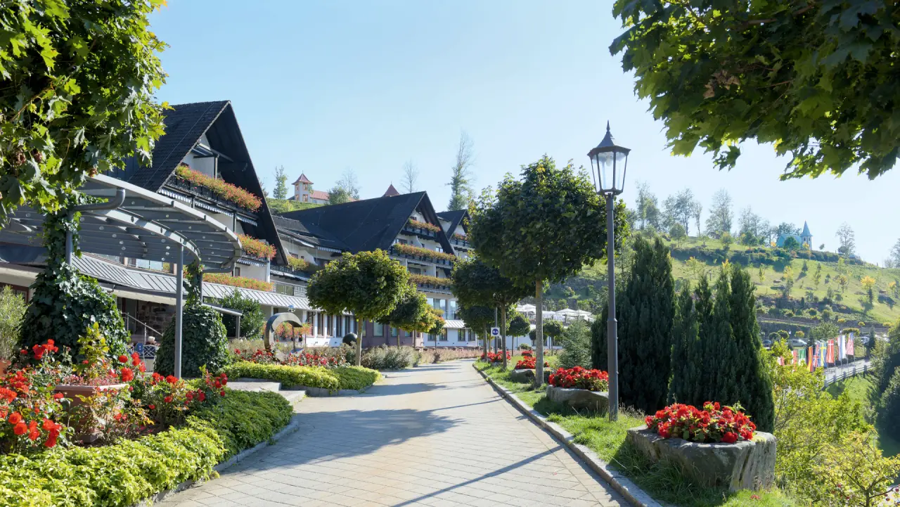 Aussenansicht des Wellnesshotels Haus Dollenberg mit gepflegtem Garten, blühenden Blumen und Zufahrtsweg unter blauem Himmel.