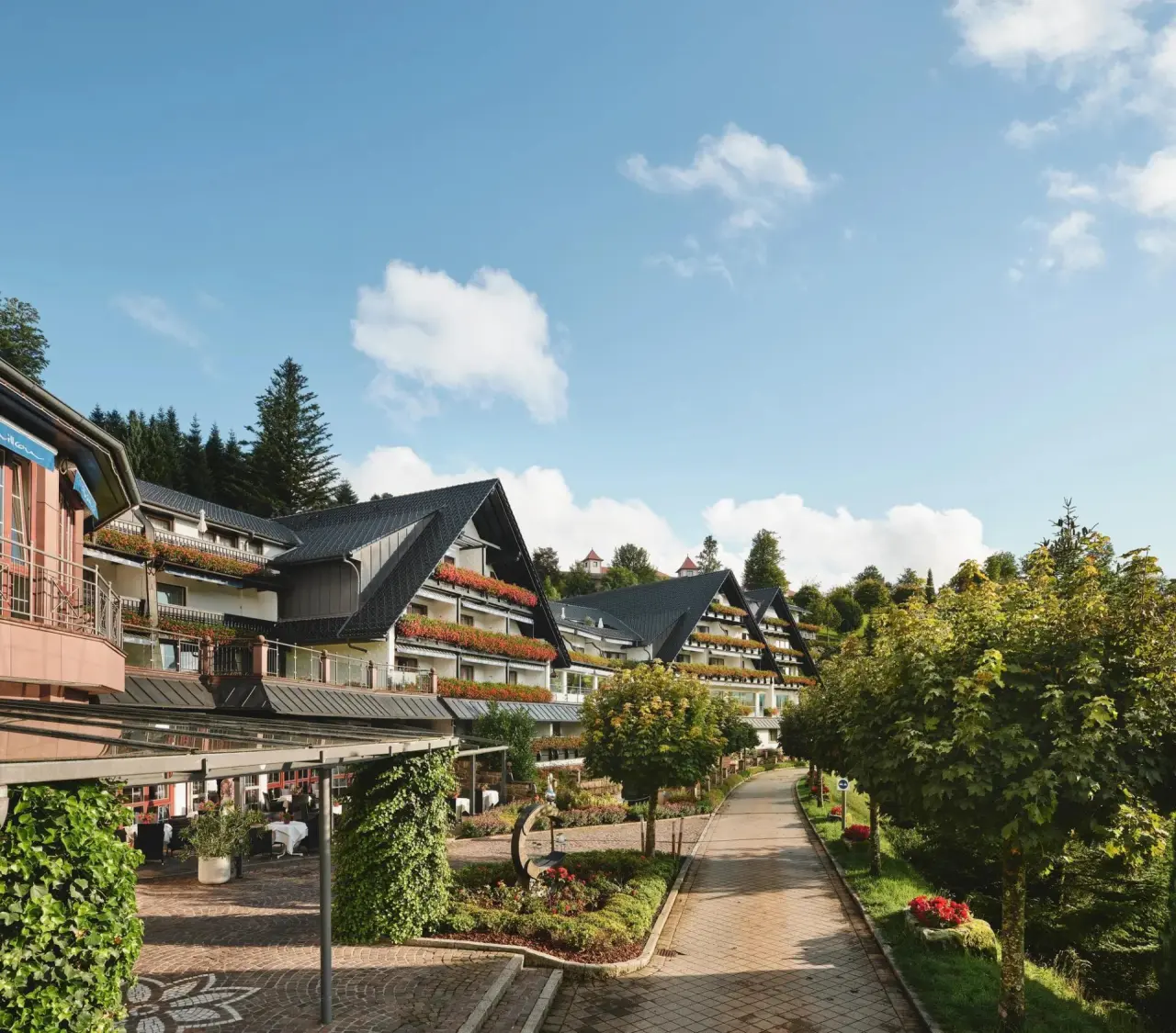Aussenansicht des Hotels Haus Dollenberg mit blühenden Balkonen, gepflegten Gärten und einem Spazierweg unter blauem Himmel.