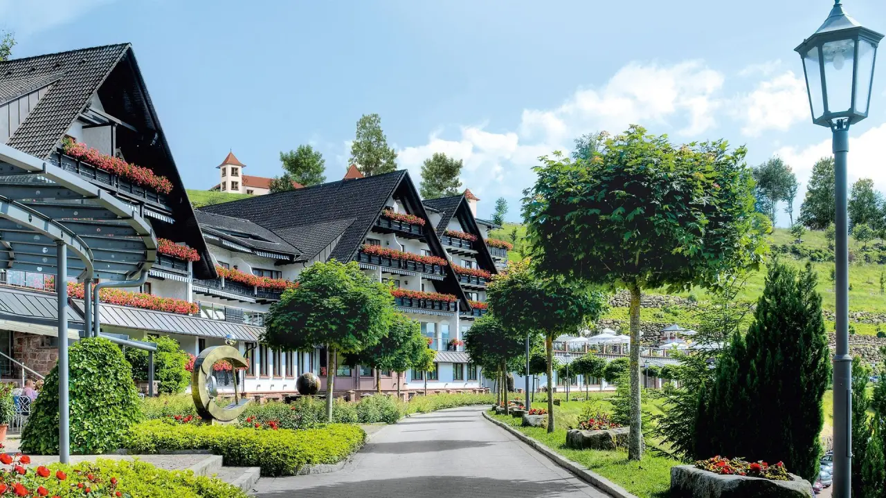 Aussenansicht des Wellness-Hotels Haus Dollenberg mit blühenden Balkonen, gepflegtem Garten und Zufahrtsweg unter blauem Himmel.