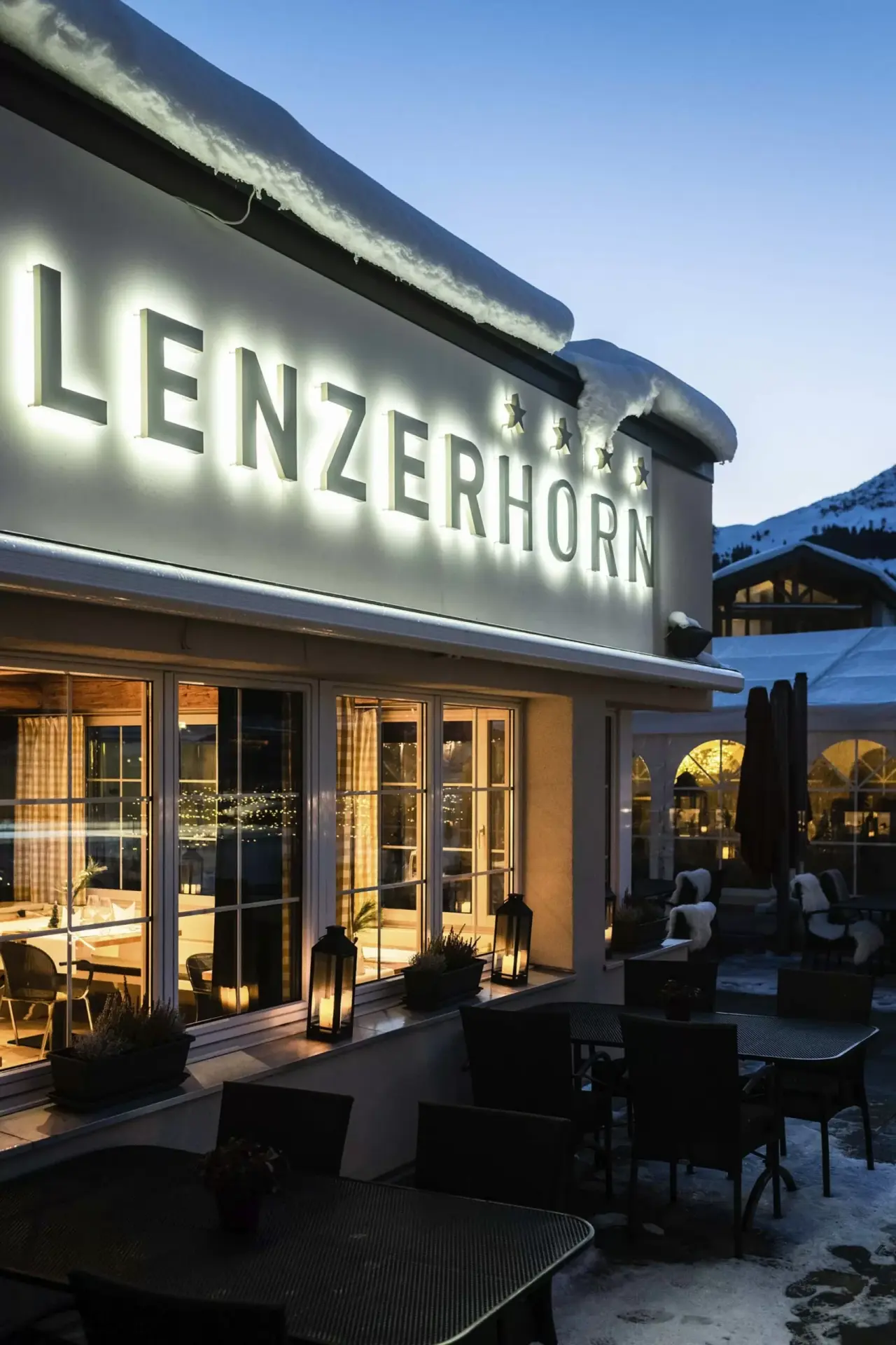 Beleuchtete Fassade des Hotel Lenzerhorn im Winter bei Dämmerung mit Schnee auf dem Dach und einladender Restaurantterrasse.