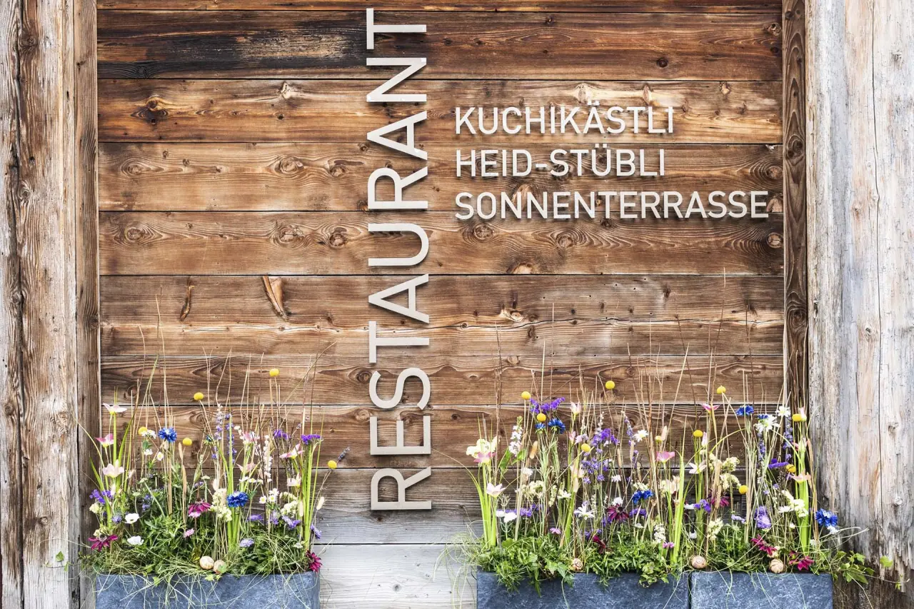 Rustikales Holzschild des Restaurants 'Kuchikästli' und 'Heid-Stübli' mit bunten Blumen am Eingang des Hotel Lenzerhorn.